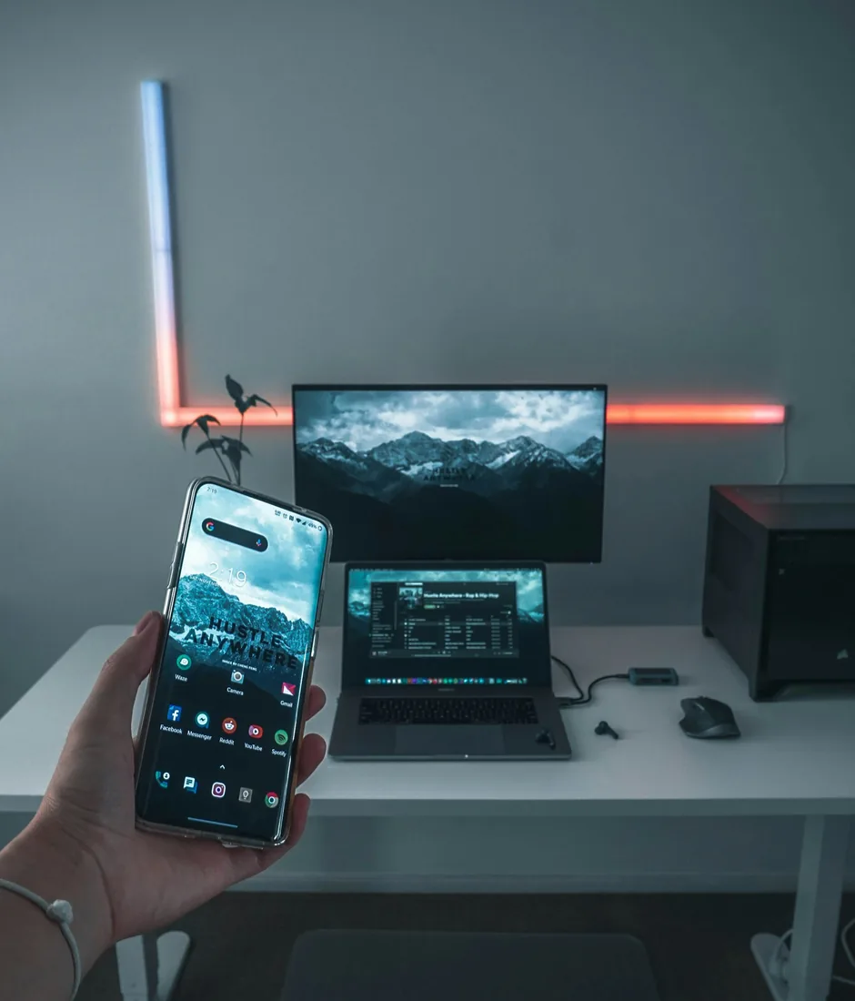 A smartphone held in front of a laptop and a monitor on a desk